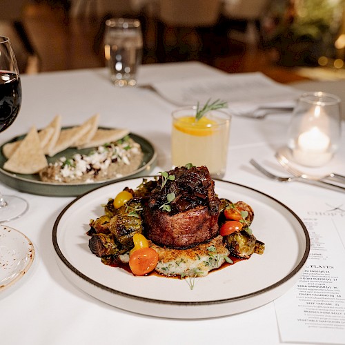 A plated steak with roasted vegetables, a glass of red wine, and other dishes on a dinner table set for a meal.