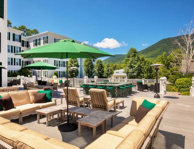 An outdoor patio with seating, umbrellas, trees, and mountains in the background on a sunny day.
