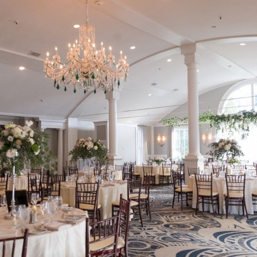 An elegant, well-lit event hall decorated for a formal occasion with round tables, floral centerpieces, and a large chandelier.
