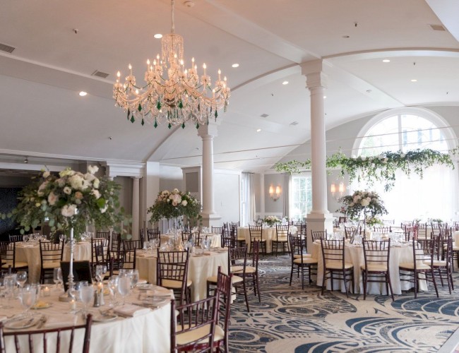 An elegant, well-lit event hall decorated for a formal occasion with round tables, floral centerpieces, and a large chandelier.