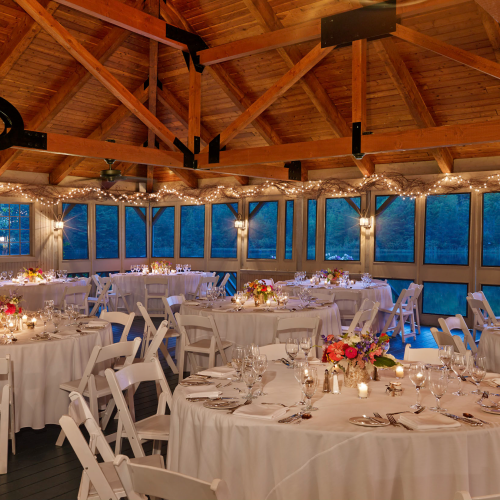 The image shows an elegantly decorated event space with round tables, white chairs, floral centerpieces, and string lights inside a wooden pavilion.