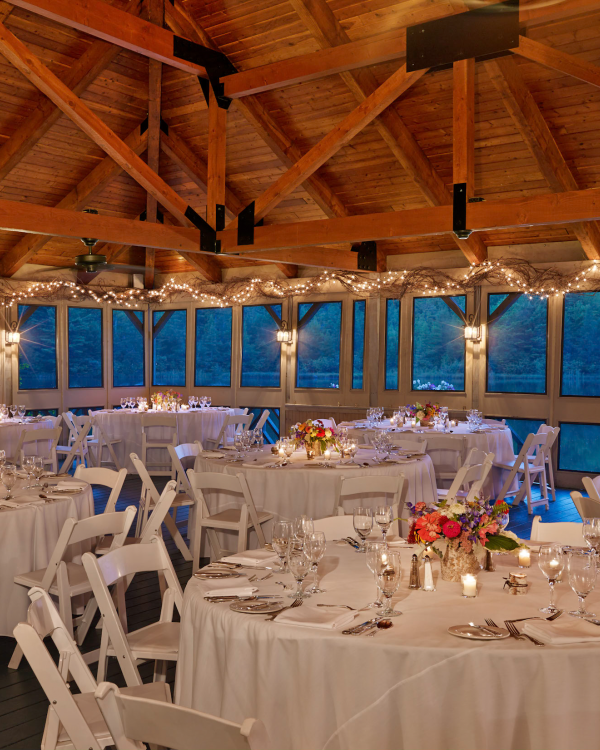 The image shows an elegantly decorated event space with round tables, white chairs, floral centerpieces, and string lights inside a wooden pavilion.