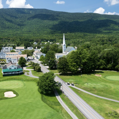 A scenic view of a small town with a golf course, church, lush trees, and a mountain in the background on a clear day.