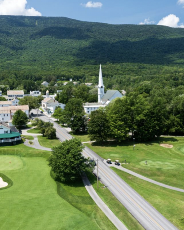 A scenic view of a small town with a golf course, church, lush trees, and a mountain in the background on a clear day.