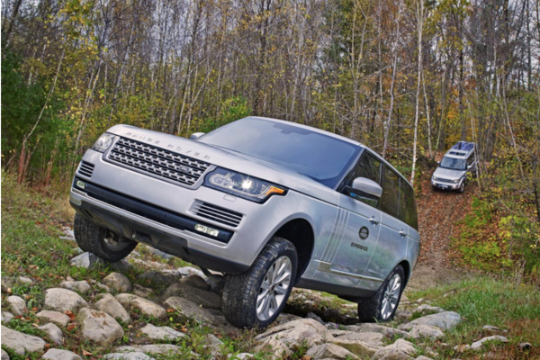 Two silver Land Rover Discovery vehicles are navigating a rocky, wooded terrain outdoors during autumn.
