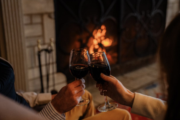 Two people are clinking glasses of red wine near a cozy fireplace in a warm, intimate setting.