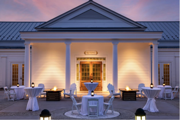 The image shows an elegant outdoor setup with tables and chairs covered in white cloth, lit by lanterns, in front of a classic white building at dusk.