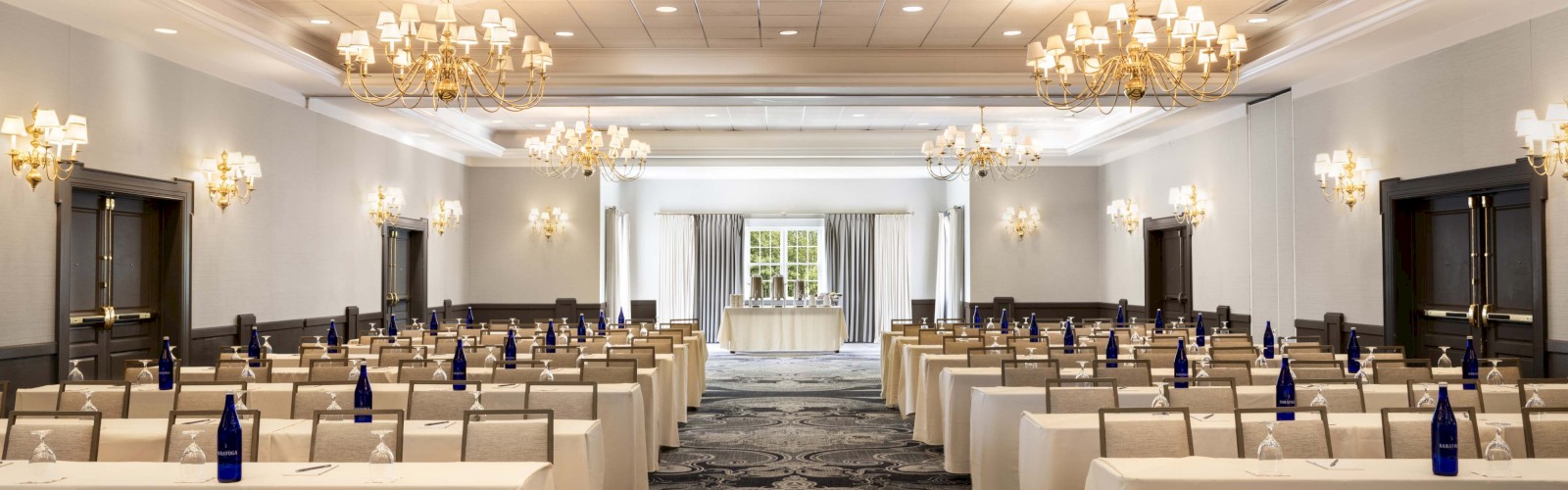 A spacious, well-lit conference or banquet room with rows of tables, each set with blue bottles and glasses, ready for an event.