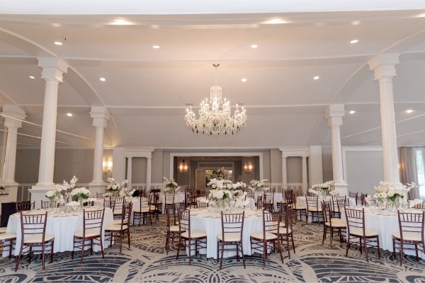 An elegant banquet hall is decorated with white tablecloths, floral centerpieces, wooden chairs, and a large chandelier.
