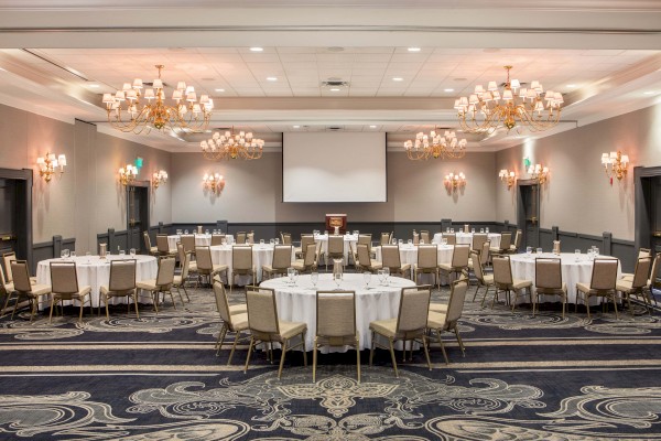 A spacious, elegant banquet hall with round tables, chandeliers, and a large projection screen at the front for events or conferences.