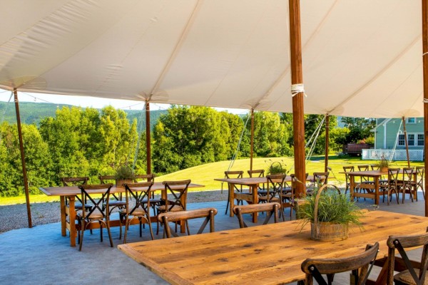 Outdoor dining setup with wooden tables and chairs under a large canopy, surrounded by greenery and a house in the background.