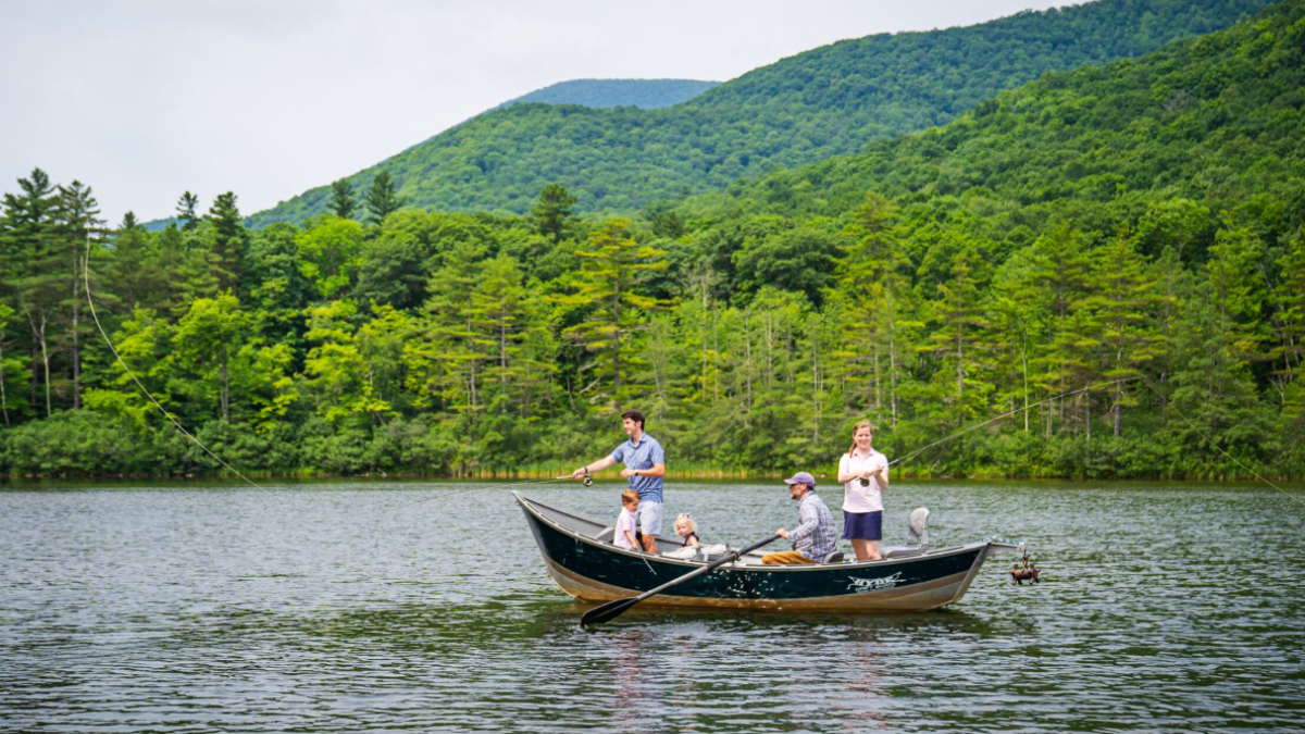 A family is fishing on a boat in a peaceful lake surrounded by green mountains and lush trees.