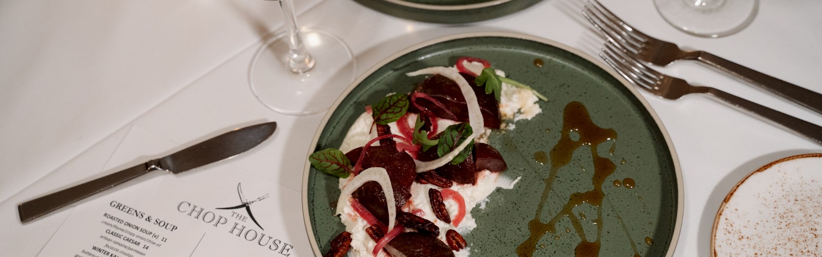 An elegant restaurant table features two plates of food, two glasses of white wine, a menu, and stainless steel cutlery.