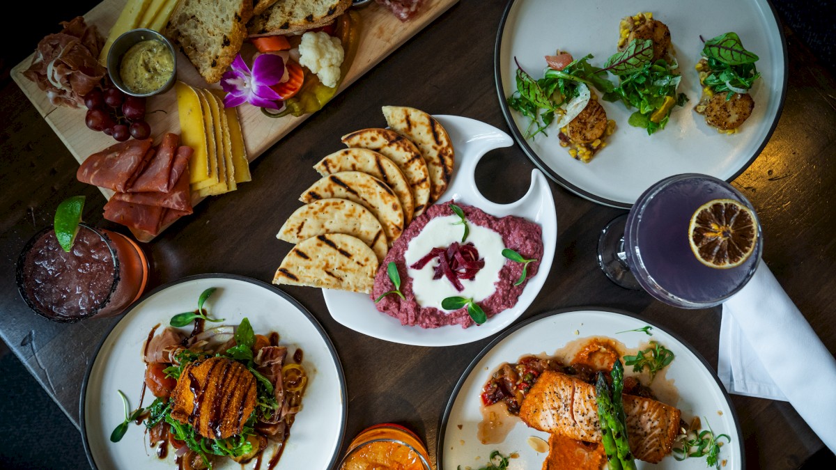 A variety of gourmet dishes and colorful beverages are displayed on a dark wooden table, featuring a charcuterie board, salads, and grilled salmon.