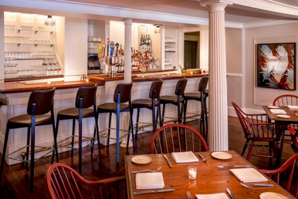 A cozy bar and restaurant with a polished countertop, bottles of alcohol, high chairs, and neatly set tables with plates, cutlery, and candles.