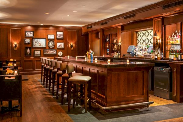A cozy bar with wooden decor, lined with high chairs, framed pictures on the wall, and shelves stocked with various bottles behind the counter.