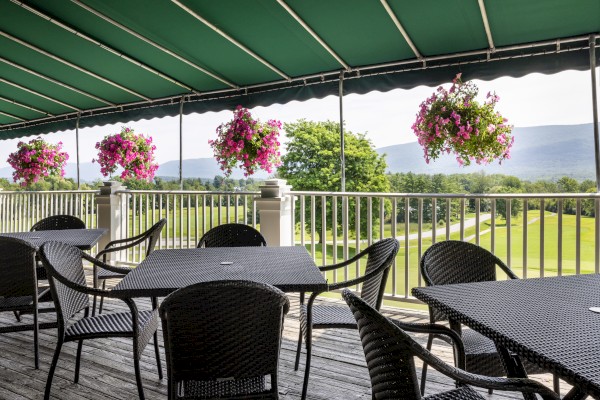 Outdoor patio with tables and chairs under a green awning, overlooking a golf course. Hanging flower baskets add a pop of color.