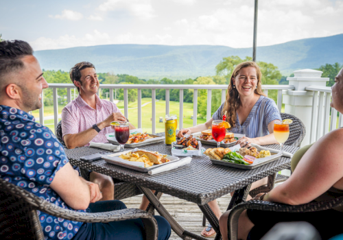 A group of friends enjoys a delicious outdoor meal on a balcony with scenic green hills and mountains in the background.
