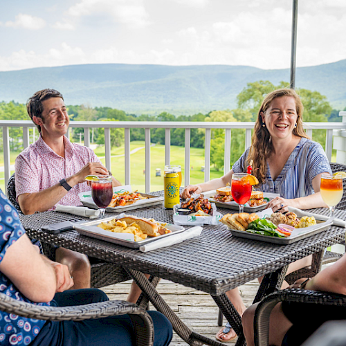 A group of friends enjoys a delicious outdoor meal on a balcony with scenic green hills and mountains in the background.