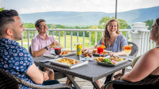 A group of friends enjoys a delicious outdoor meal on a balcony with scenic green hills and mountains in the background.