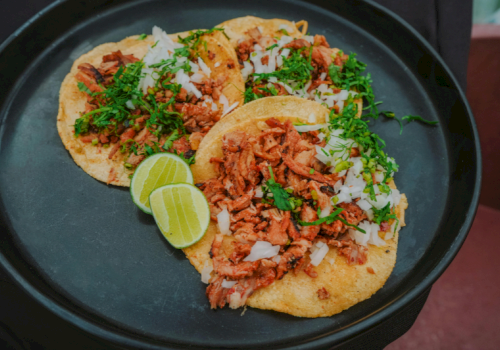 Three tacos filled with shredded meat, topped with herbs and chopped onions, with lime wedges on a black plate.