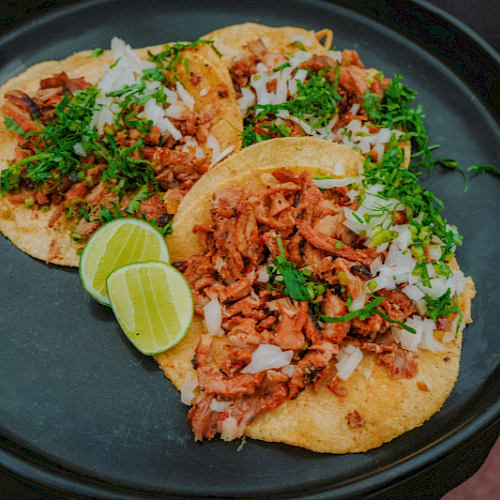 Three tacos filled with shredded meat, topped with herbs and chopped onions, with lime wedges on a black plate.