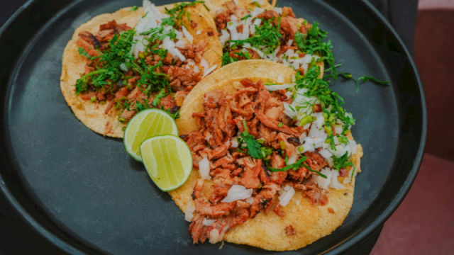 Three tacos filled with shredded meat, topped with herbs and chopped onions, with lime wedges on a black plate.