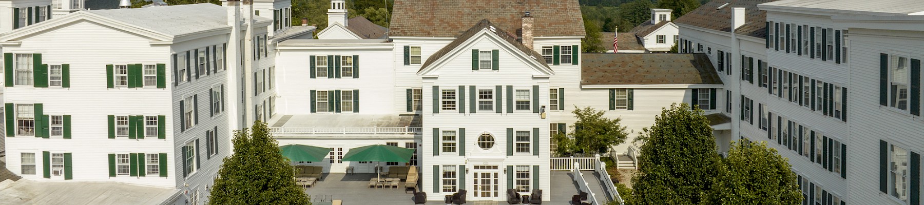 This image shows a charming white building with green shutters, a terrace with umbrellas, and a backdrop of green mountains.