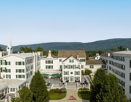 This image shows a charming white building with green shutters, a terrace with umbrellas, and a backdrop of green mountains.