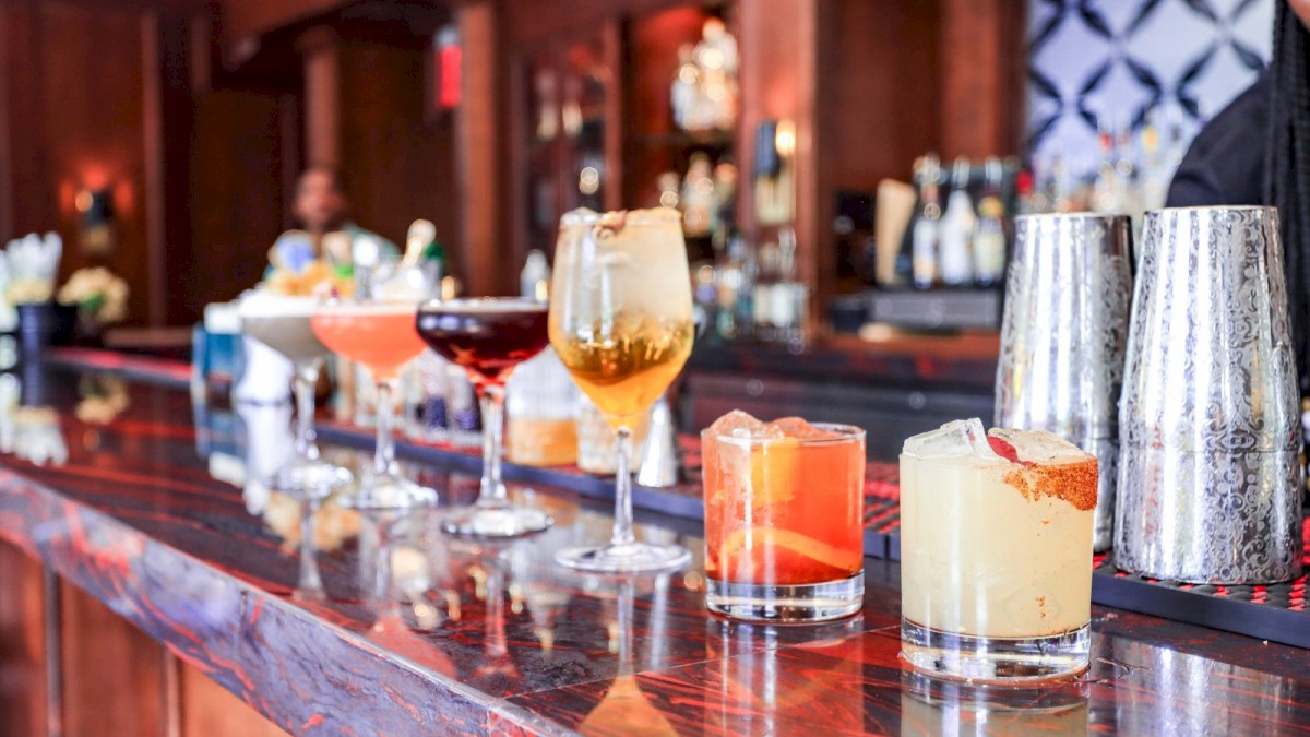 The image shows a variety of colorful cocktails and drinks on a polished wooden bar counter in a cozy, well-lit bar setting with a blurred bartender in the background.