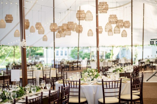 This image shows an elegantly decorated outdoor wedding reception under a tent with round tables, white tablecloths, floral centerpieces, and hanging lanterns.