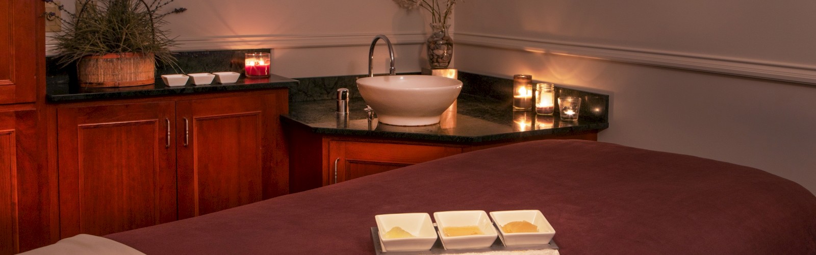 A cozy massage or spa room with candles, herbal decor, a wash basin, and three bowls of oil or lotion for relaxation.