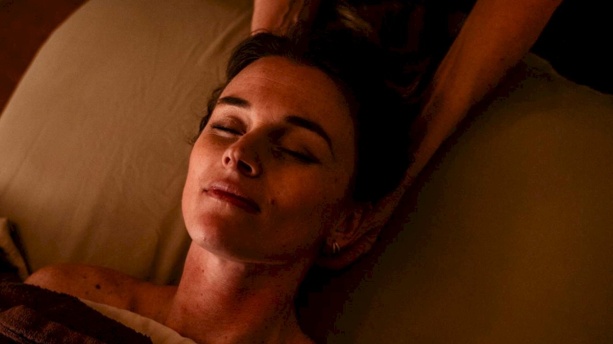 A woman with closed eyes relaxes as a person gives her a head massage on a bed in dim lighting.