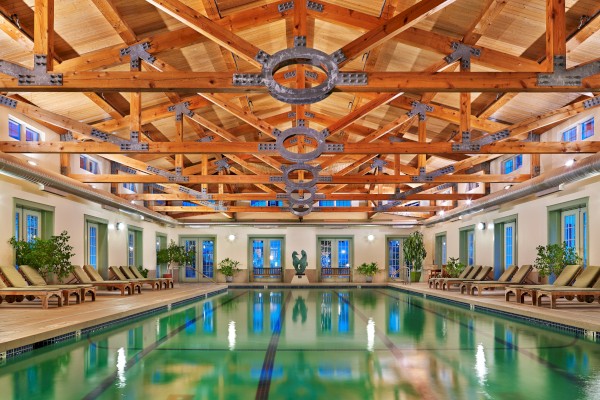 This is an indoor swimming pool area with wooden ceiling beams, multiple windows, plants, and lounge chairs along the sides.