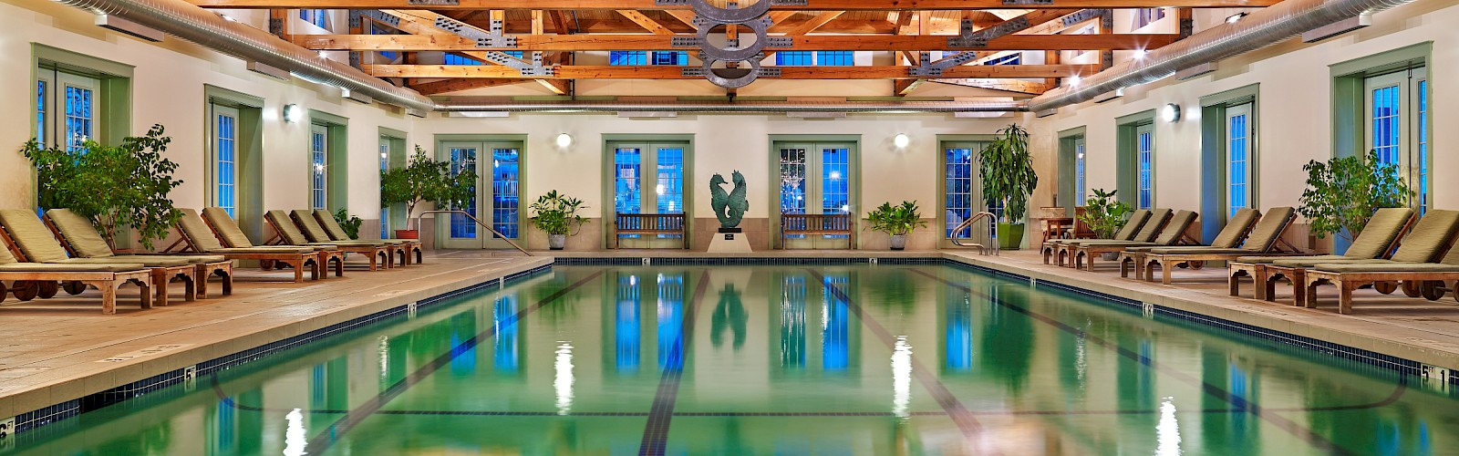 This is an indoor swimming pool with wooden ceiling beams, potted plants, lounge chairs, and a decorative sculpture near the back.