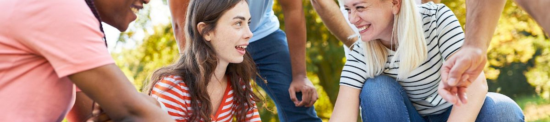 A diverse group of friends is outdoors, smiling and pointing at a map on the grass during a sunny day.