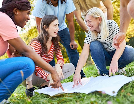 A diverse group of friends is outdoors, smiling and pointing at a map on the grass during a sunny day.