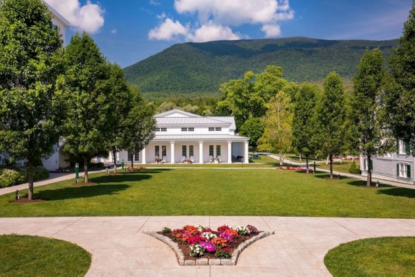 A scenic view of a white building surrounded by green trees and a mountain in the background, with colorful flowers and well-maintained lawns.