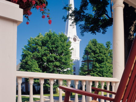 A porch view showing a red rocking chair, pink flowers, green trees, and a white church steeple against a blue sky.