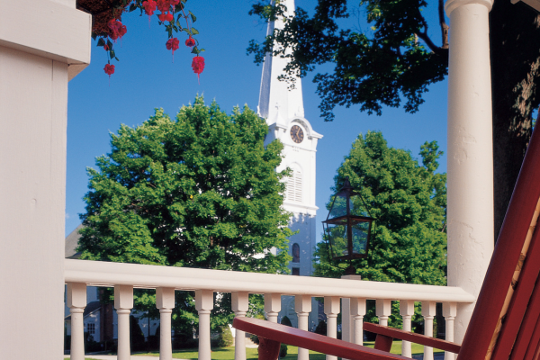 A porch view showing a red rocking chair, pink flowers, green trees, and a white church steeple against a blue sky.
