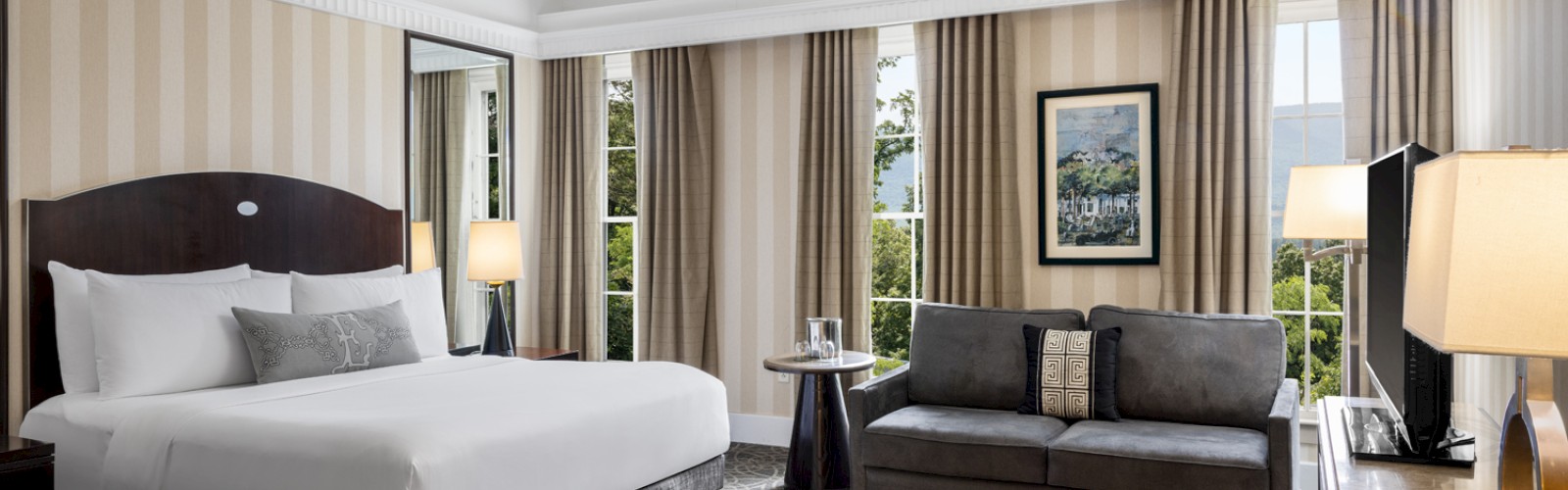 A luxurious hotel room with a large bed, a gray sofa, multiple windows with curtains, a TV, lamps, and art on the wall.