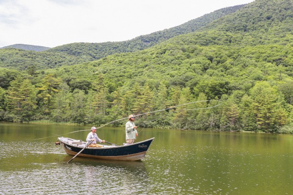 Two men fish from a boat on a peaceful lake surrounded by lush green mountains and trees.