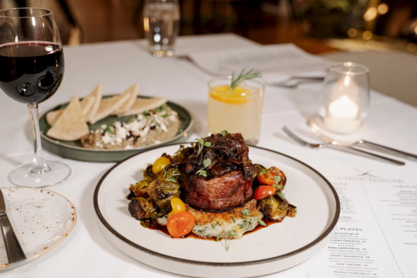 A table with a glass of red wine, a plated main dish, a drink, a candle, and a menu at a fancy restaurant dinner.