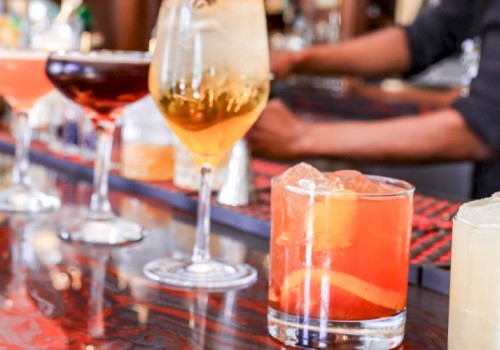 A row of colorful cocktails and drinks lined up on a bar counter, with bartenders and bottles in the background.