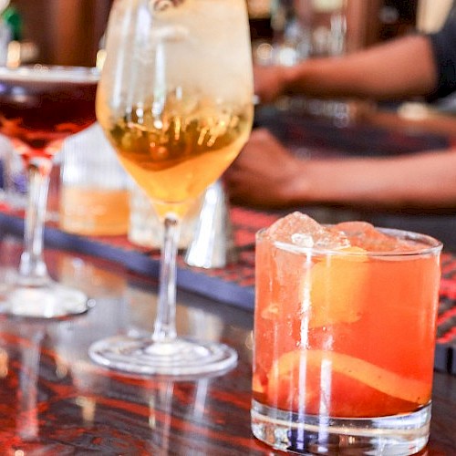A row of colorful cocktails and drinks lined up on a bar counter, with bartenders and bottles in the background.