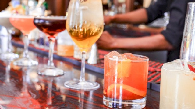 A row of colorful cocktails and drinks lined up on a bar counter, with bartenders and bottles in the background.