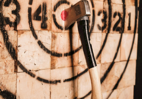 An axe embedded in a painted wood target, likely for axe throwing, with concentric circles numbered for scoring.