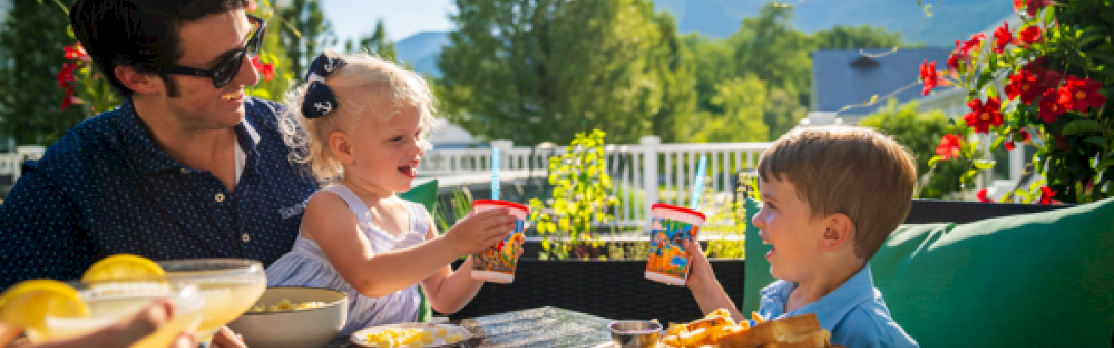 A man and two children enjoy snacks and drinks on a sunny outdoor patio with colorful flowers and mountains in the background.
