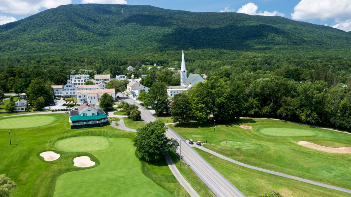 A scenic village with a church, surrounded by lush greenery, mountains, and a golf course with several sand bunkers.