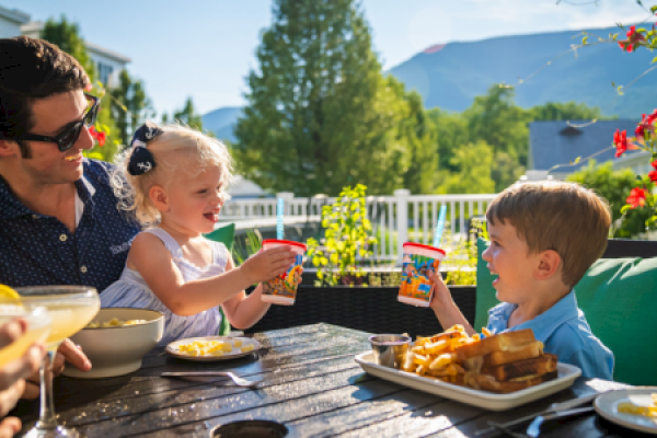 A family is enjoying a sunny outdoor meal with drinks, snacks, and a cheerful moment between a woman, a girl, and a boy.